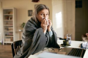 Imagem mostra mulher grisalha sentada em uma mesa, com uma coberta e assoando o nariz - Metrópoles
