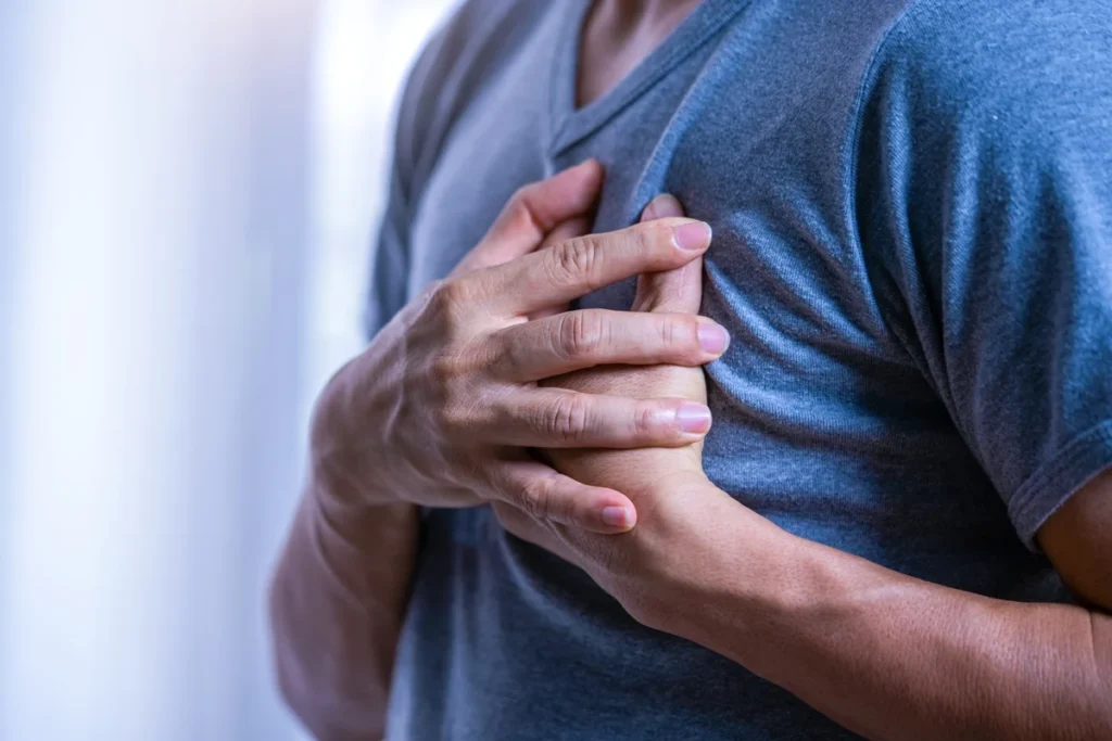 Foto colorida de um homem com blusa azul e mãos no peito, com sintomas de infarto - Metrópoles.