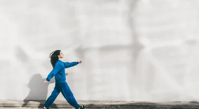 Mulher jovem de roupa azul caminhando em frente à parede branca - Metrópoles