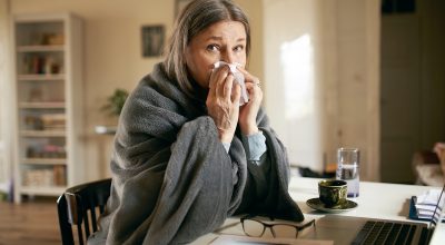 Imagem mostra mulher grisalha sentada em uma mesa, com uma coberta e assoando o nariz - Metrópoles