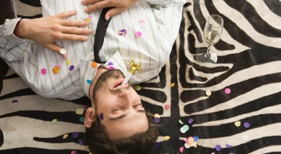 Foto colorida de homem coberto por confeti deitado sobre um tapete ao lado de uma taça com bebida - Metrópoles.