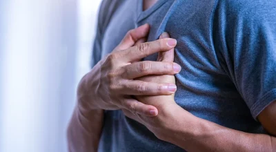 Foto colorida de um homem com blusa azul e mãos no peito, com sintomas de infarto - Metrópoles.